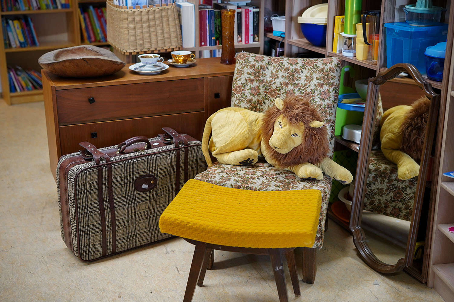 Ein alter Stuhl mit Blumenbezug auf dem ein Kuscheltier Löwe sitzt. Dahinter sind verschiedene gebrauchte Möbel und ein Stoff-Koffer zu sehen.